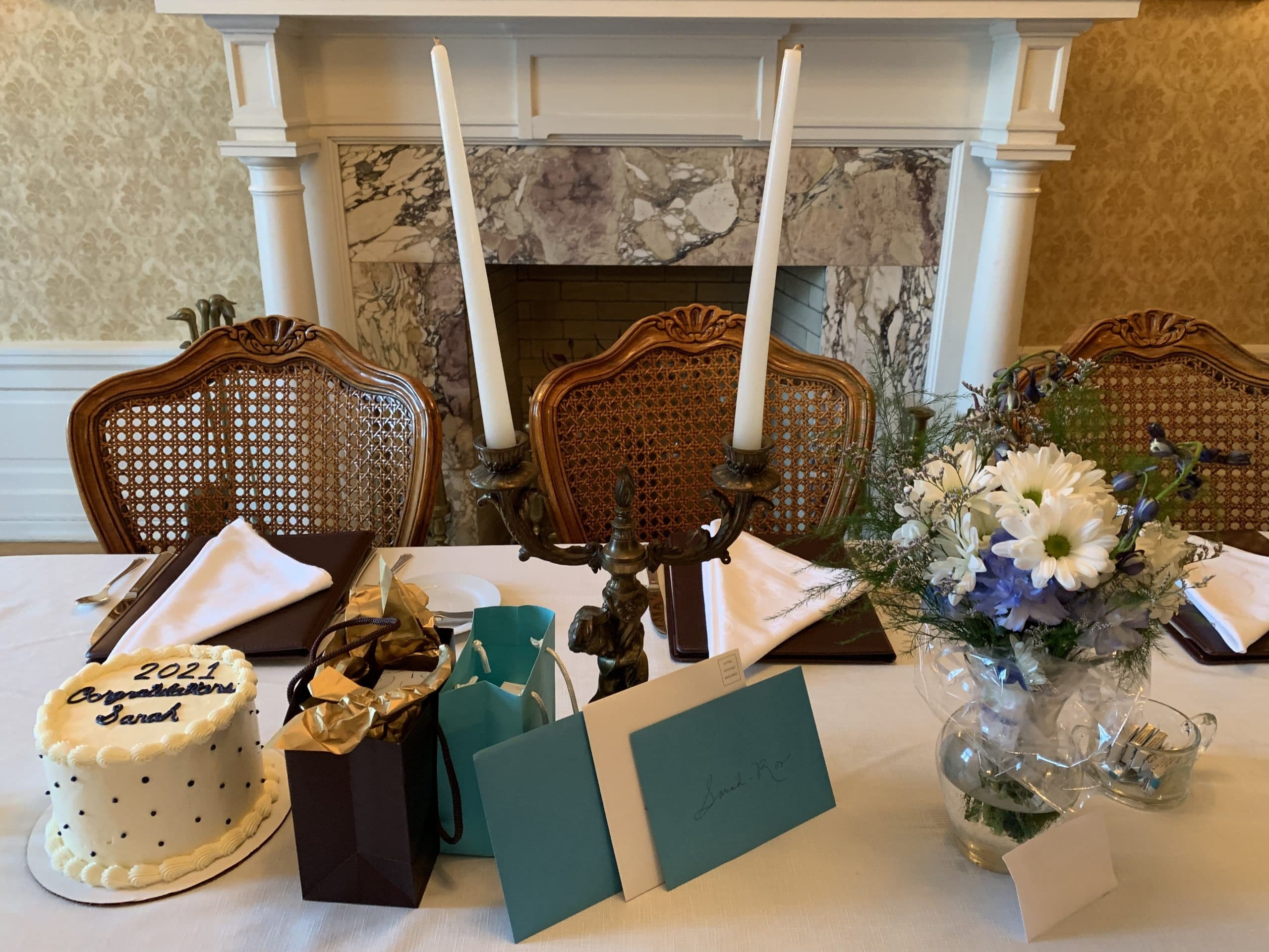 A decorated table featuring a cake, gifts, cards, and a flower arrangement, with a fireplace in the background.