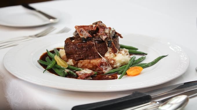 A plated beef filet topped with bacon, served with mashed potatoes and green beans.