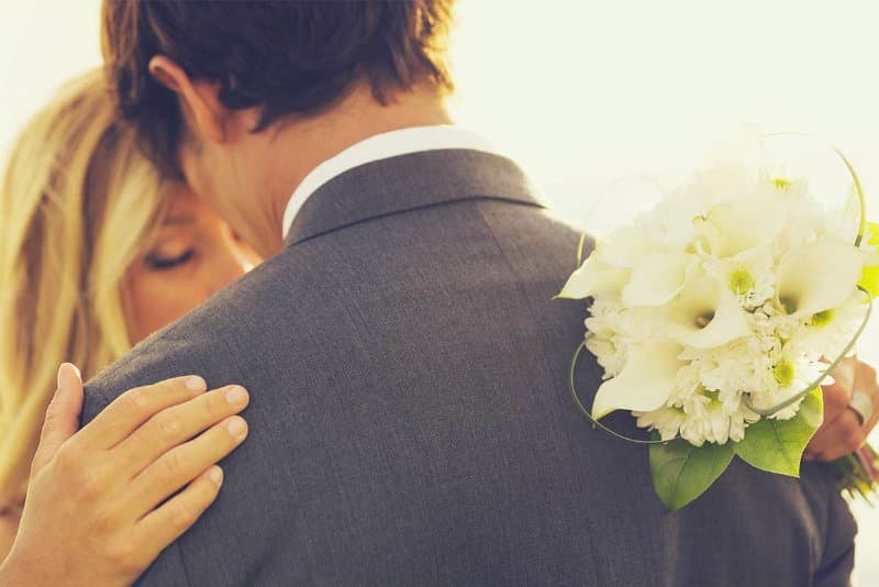 A couple embraces, with a bouquet of white flowers held against the man's shoulder.