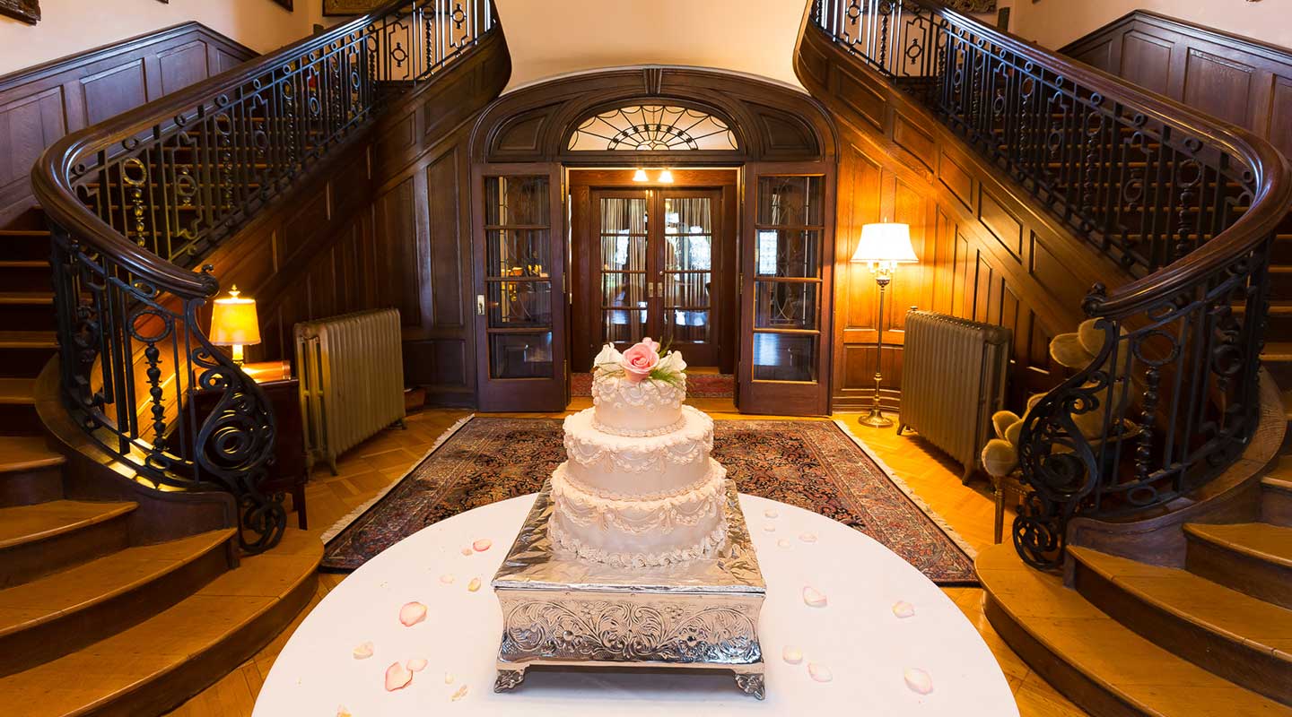 A decorated wedding cake stands on a white table in a grand entrance hall with a wooden staircase.