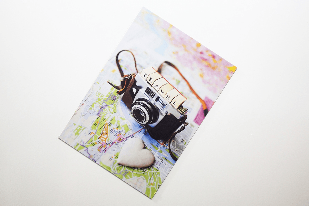 A vintage camera rests on a colorful map with a heart-shaped stone, accompanied by the word "TRAVEL."