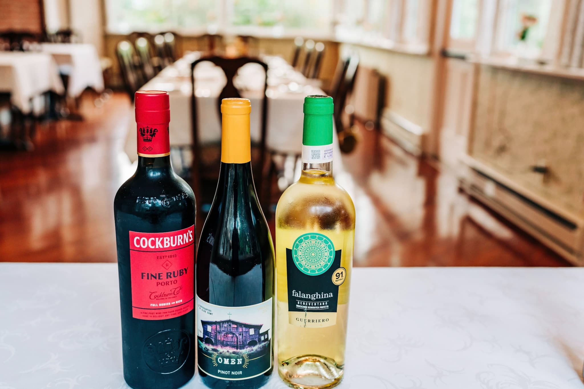 Three bottles of wine on a table: a red port, a Pinot Noir, and a white Falanghina, with a dining area in the background.