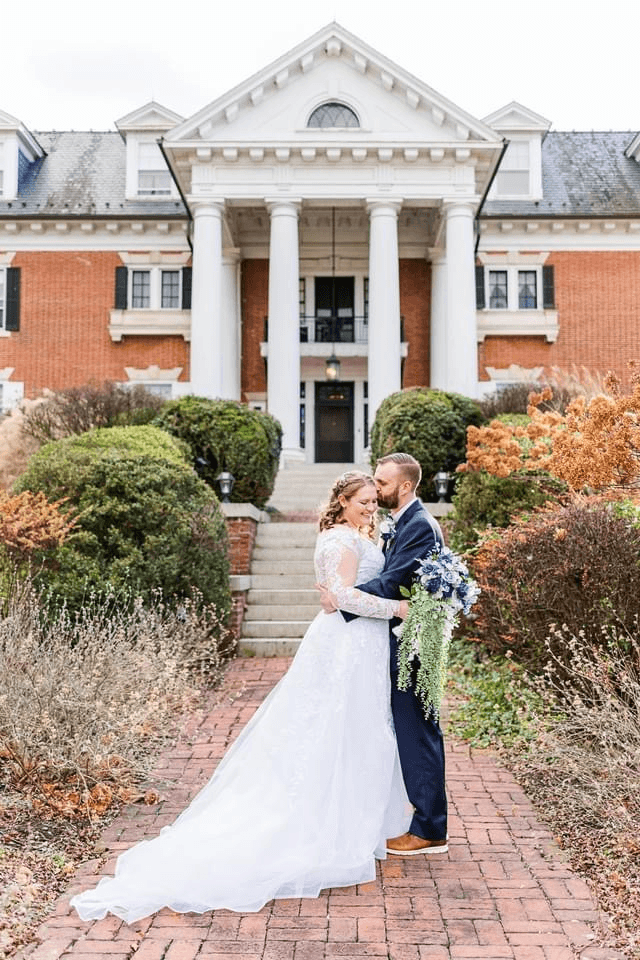 A couple embraces on a brick path in front of a grand building, adorned with lush greenery and autumn foliage.