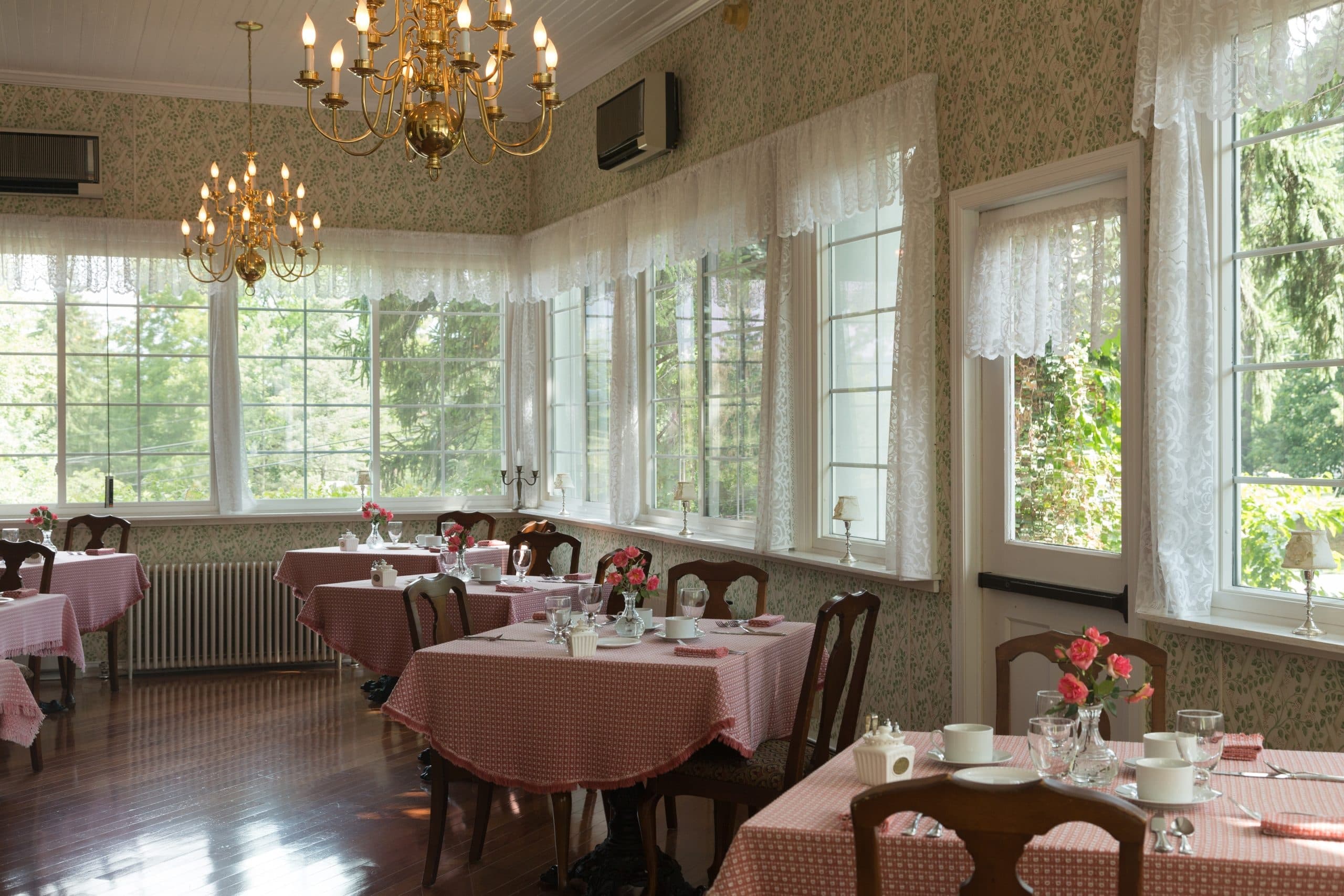 A cozy dining room with pink checkered tablecloths, floral centerpieces, and large windows letting in natural light.