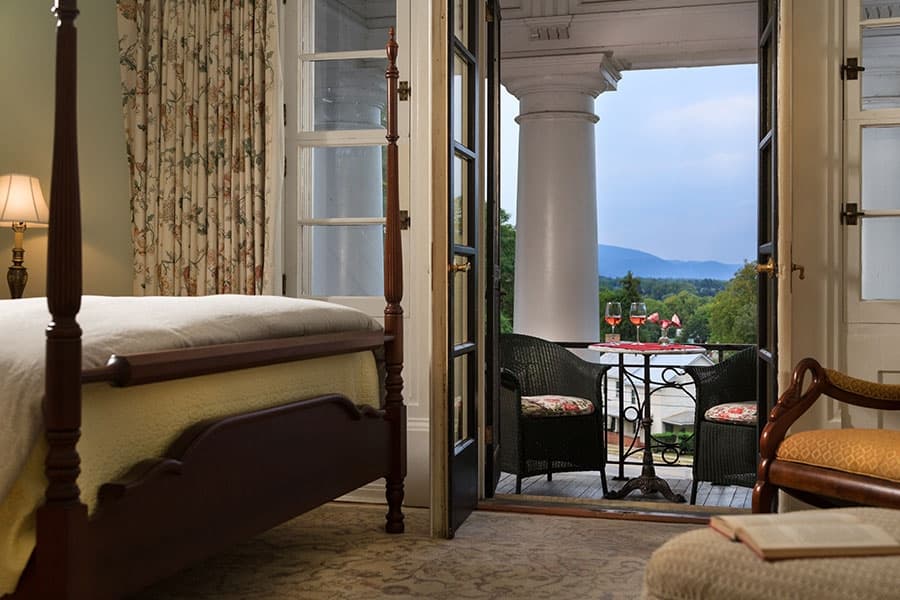 A cozy bedroom with a view of a balcony and mountains beyond, featuring a four-poster bed and floral curtains.