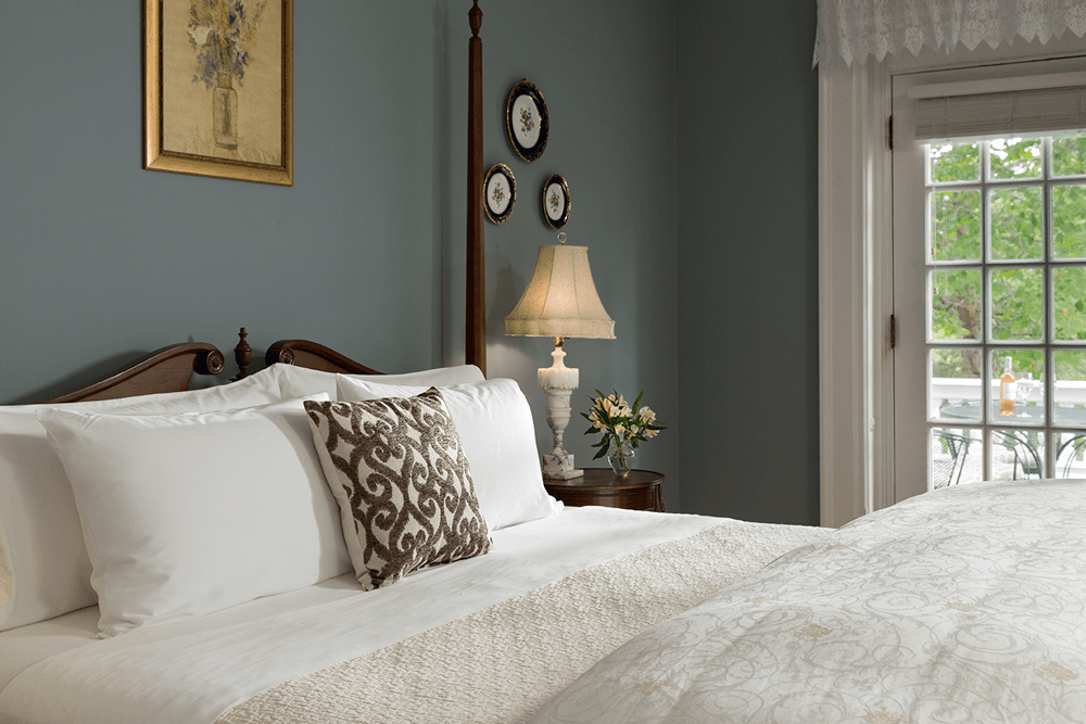 A cozy bedroom featuring a wooden bed with decorative pillows, a lamp, and floral artwork against a blue wall.