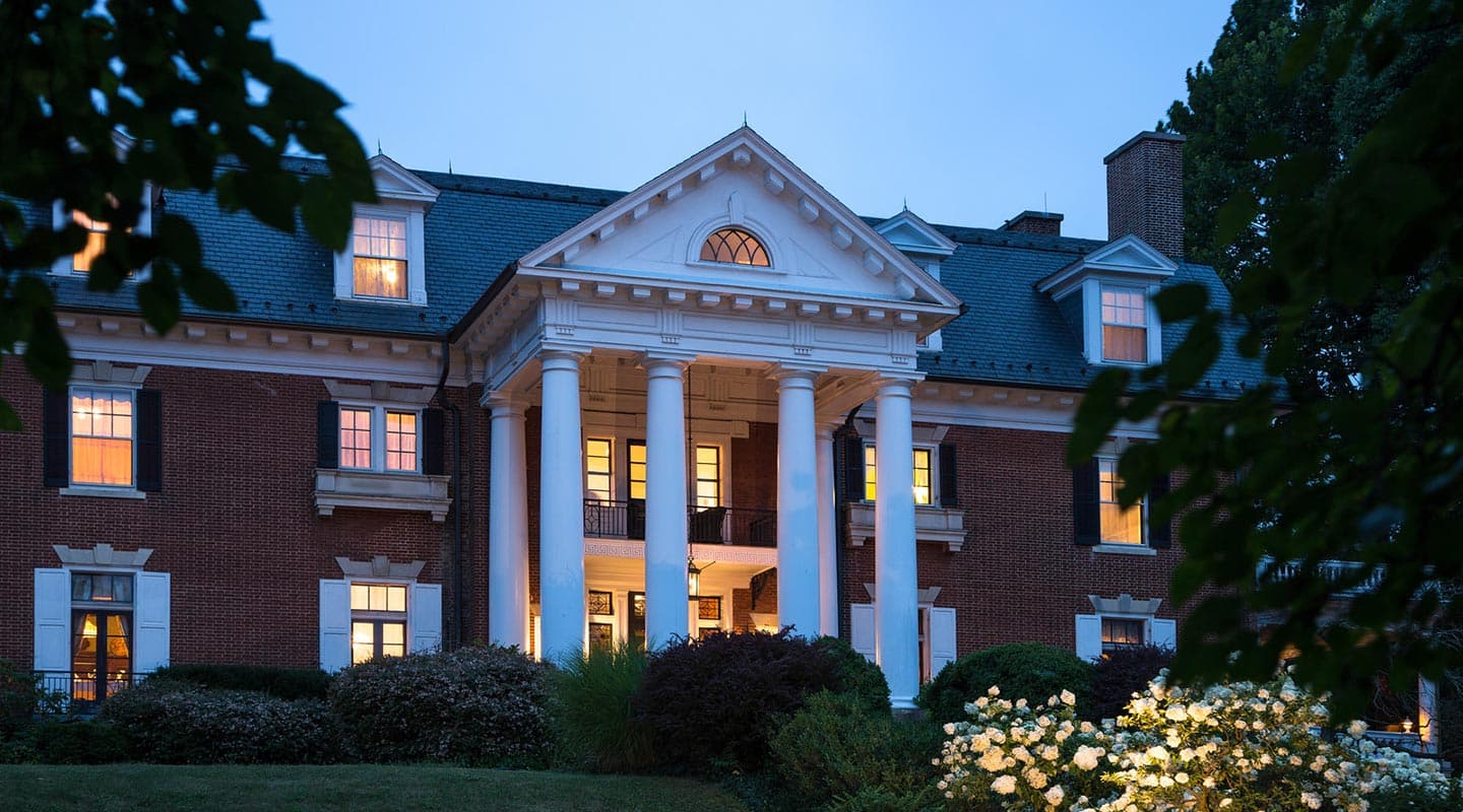 A grand brick mansion with white columns and illuminated windows surrounded by greenery at dusk.