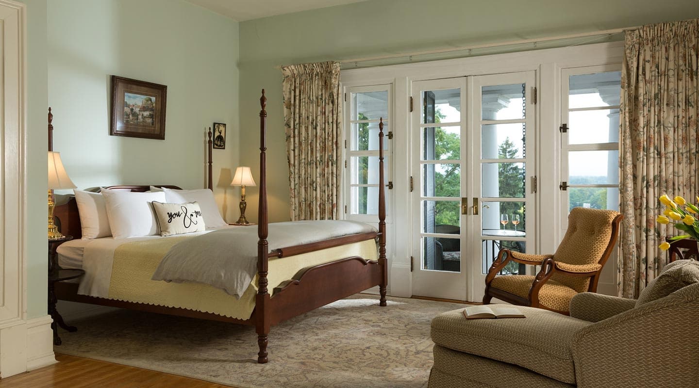 A cozy, well-lit bedroom featuring a four-poster bed, a comfortable armchair, and large French doors opening to a scenic view.