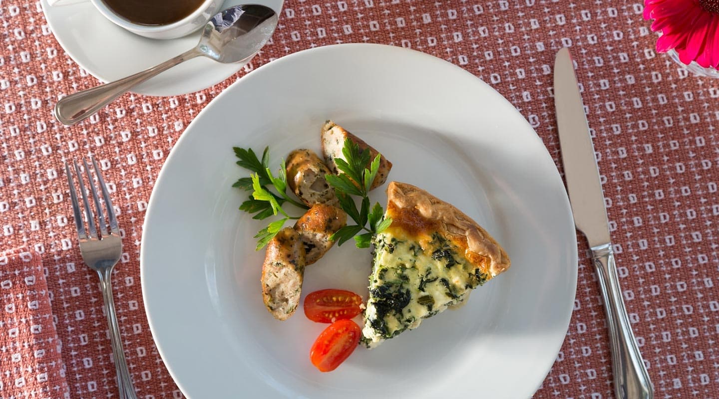 A plate featuring a slice of spinach quiche, sausage, cherry tomatoes, and a garnish of parsley alongside a cup of coffee.