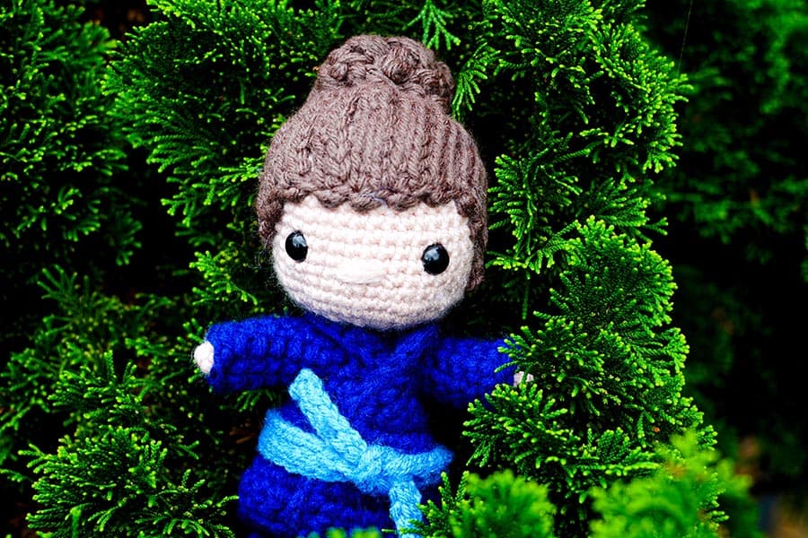 A crocheted doll in a blue outfit with a brown bun hairstyle nestled among green foliage.