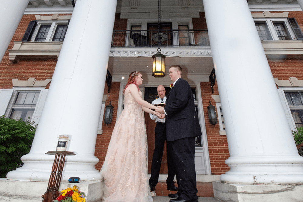 A bride and groom exchange vows under a lantern at a grand entrance, while an officiant oversees the ceremony.