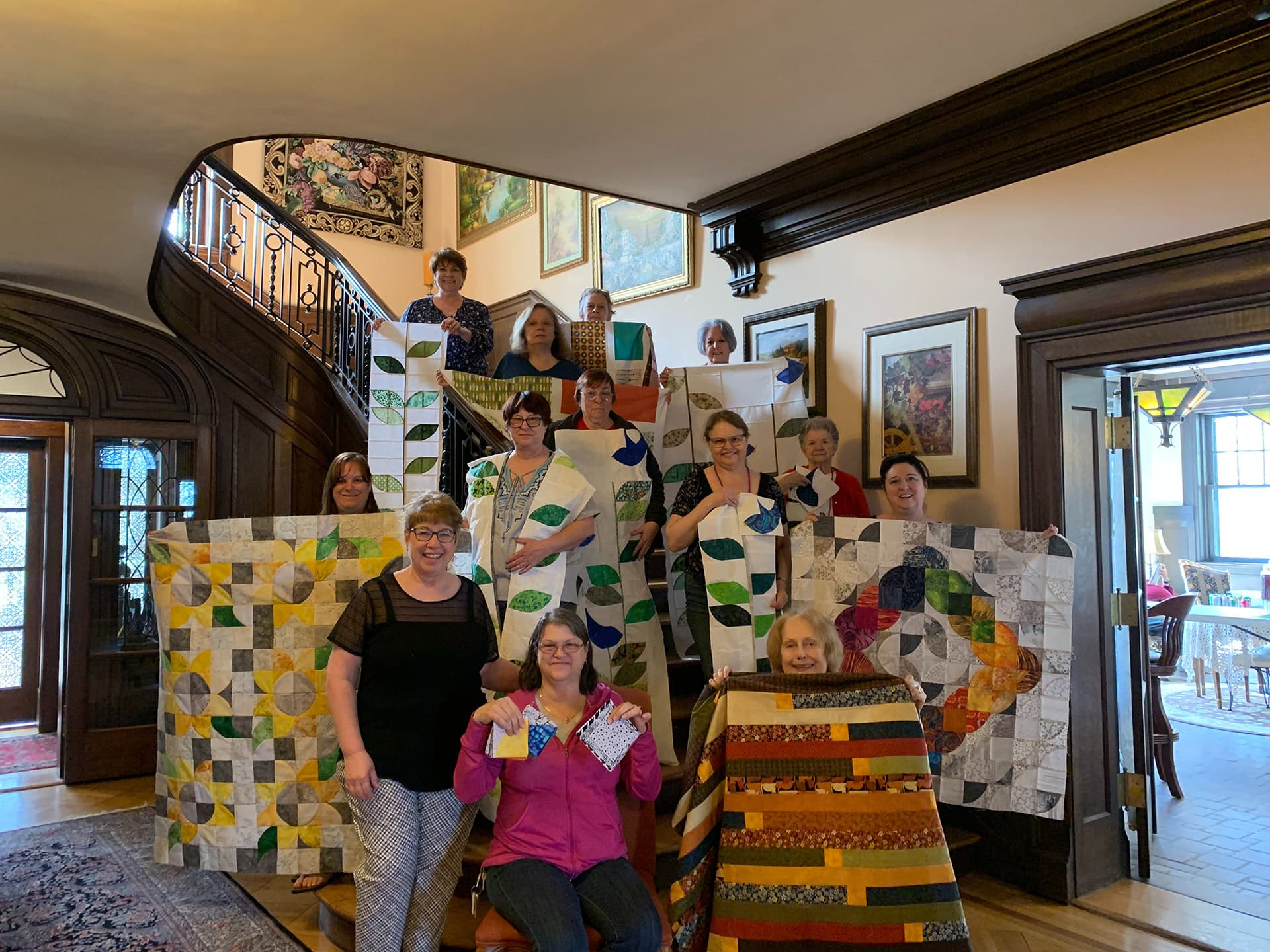 A group of quilters stands on a staircase, proudly displaying their colorful quilt creations.