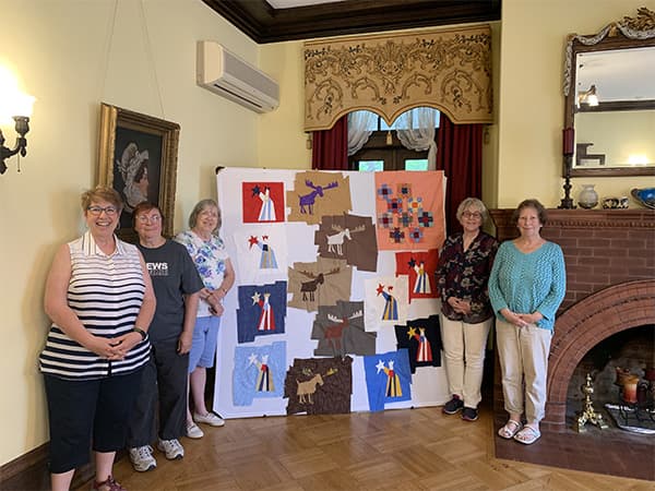 A group of six women stands beside a colorful quilt display in a warmly decorated room.