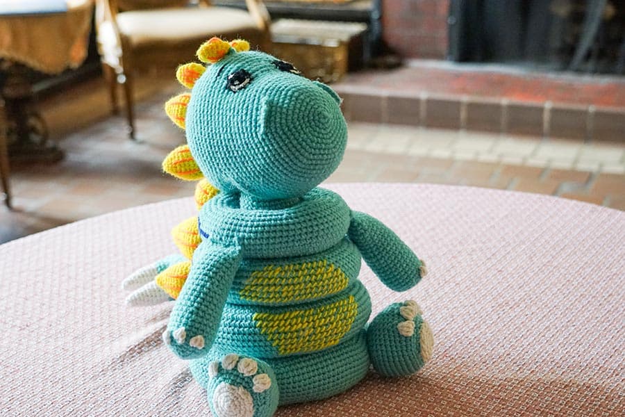 A handmade, crocheted dragon toy in teal with yellow accents, sitting on a table.