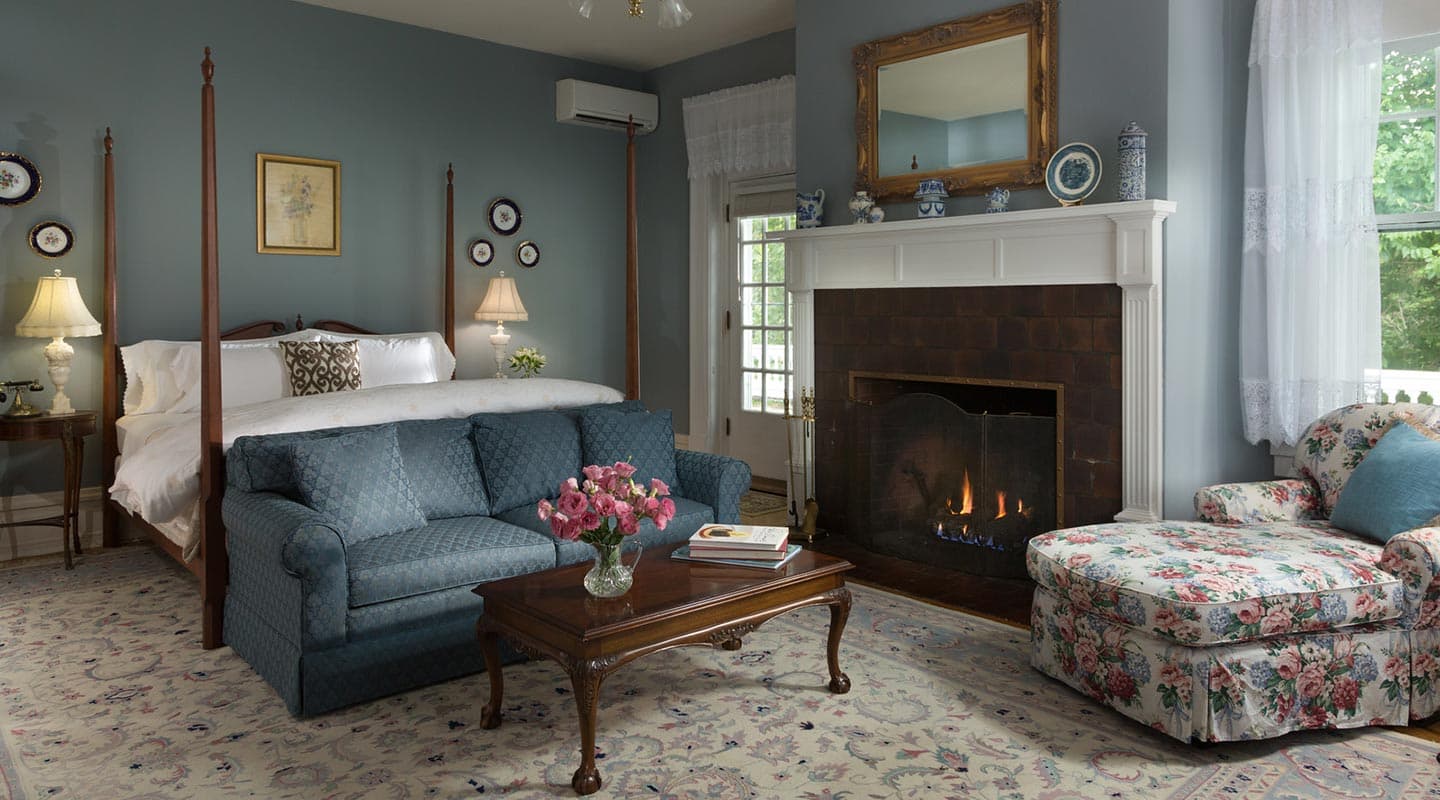 Cozy bedroom featuring a four-poster bed, fireplace, floral armchair, and blue sofa with a coffee table.