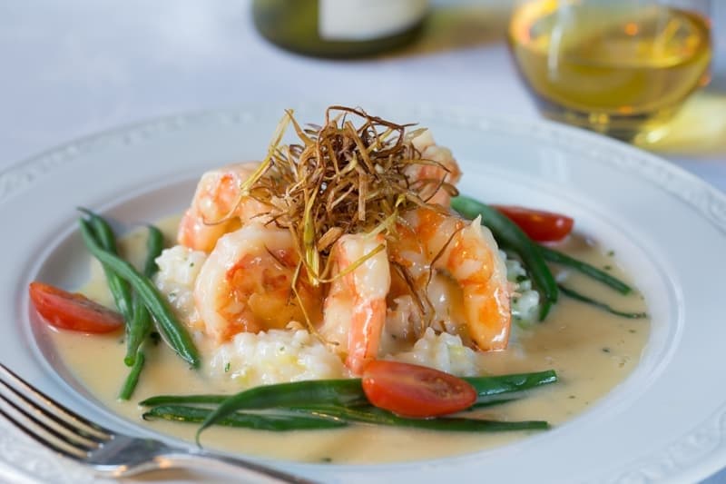 A plate of shrimp atop creamy risotto, garnished with green beans, cherry tomatoes, and crispy fried shallots.
