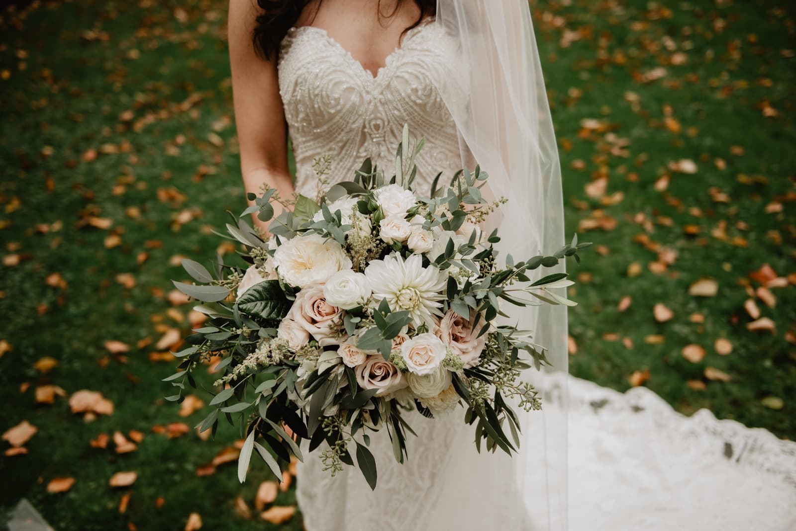 A bride in a lace wedding gown holds a bouquet of cream and blush roses with greenery.