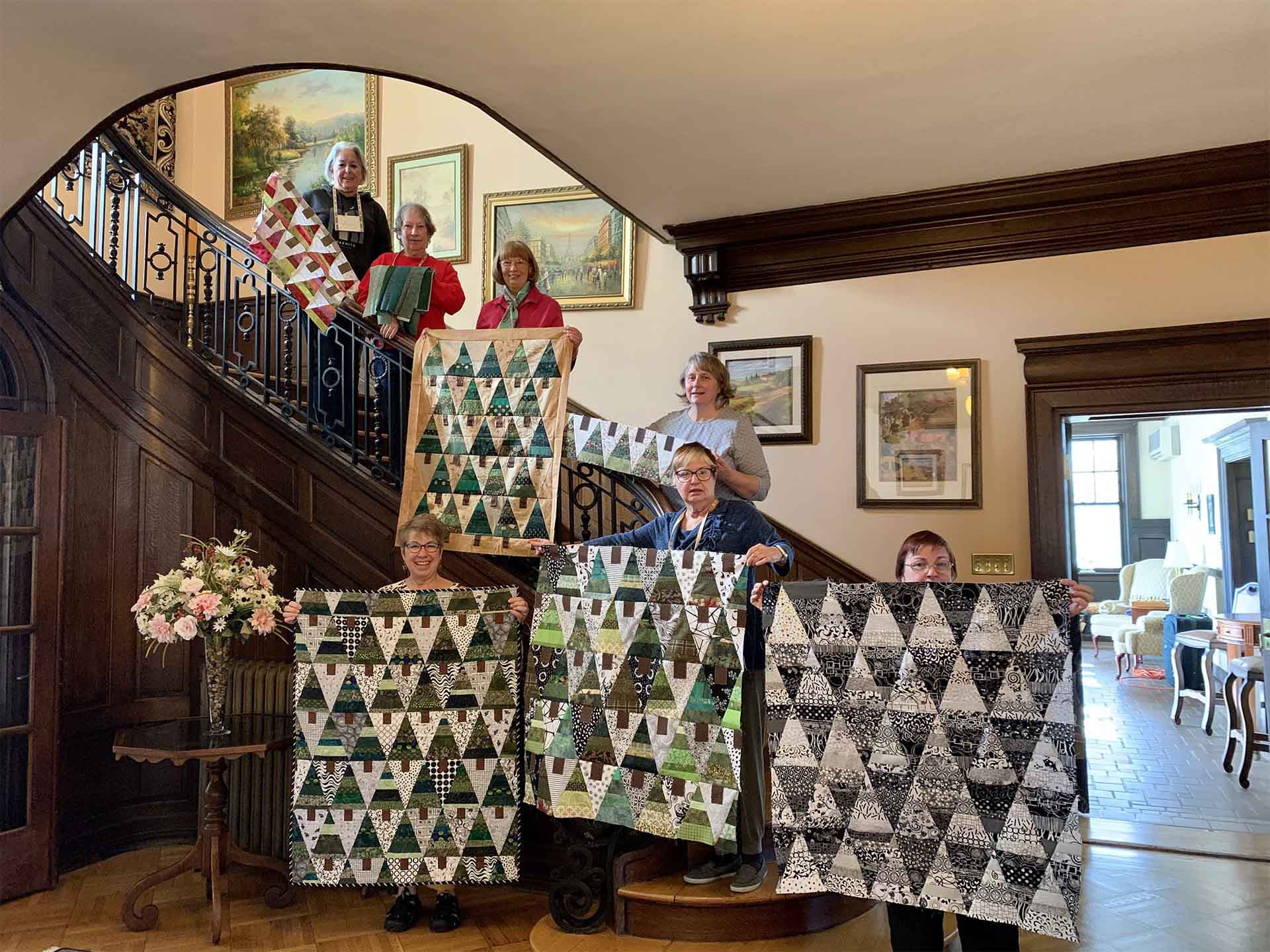 A group of seven women poses on a staircase, each holding colorful quilts with triangular patterns.