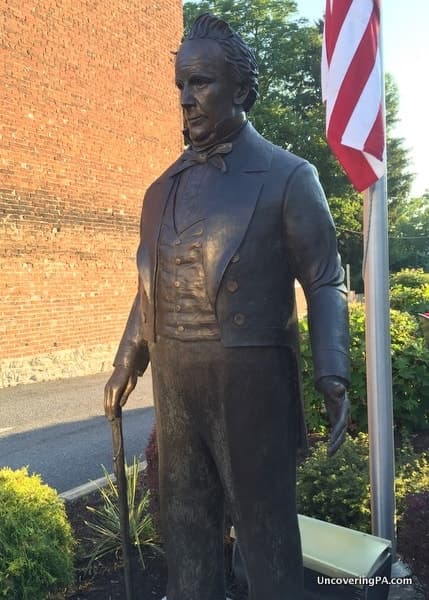 A bronze statue of a well-dressed man holding a cane, with an American flag in the background.