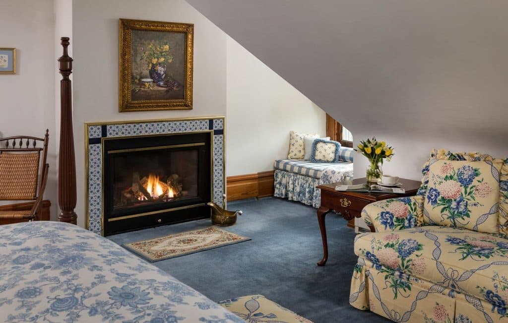 A cozy attic room featuring a fireplace, floral-patterned furniture, and a view of a window seat.