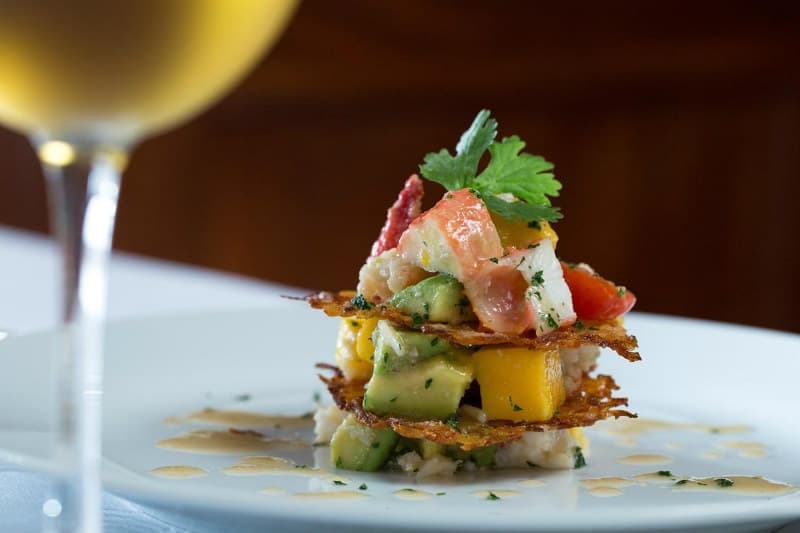 A colorful stacked dish featuring seafood, avocado, and mango on a white plate, accompanied by a glass of wine.