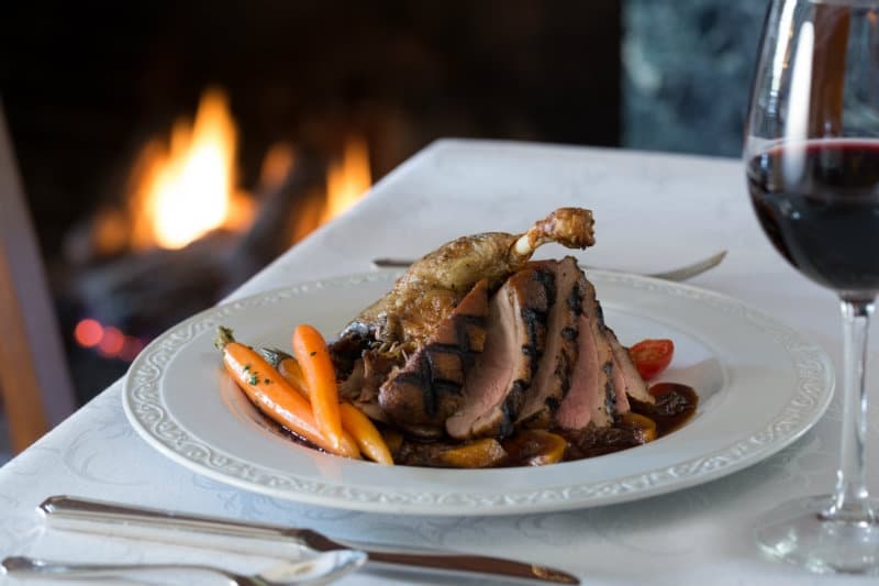 A gourmet plate of sliced duck breast and roasted vegetables, accompanied by a glass of red wine and a warm fireplace in the background.
