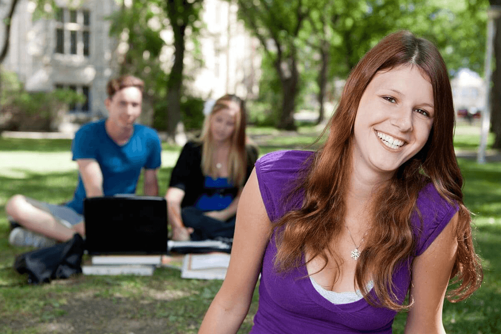 A smiling young woman in a purple shirt sits in a park with two classmates working behind her.