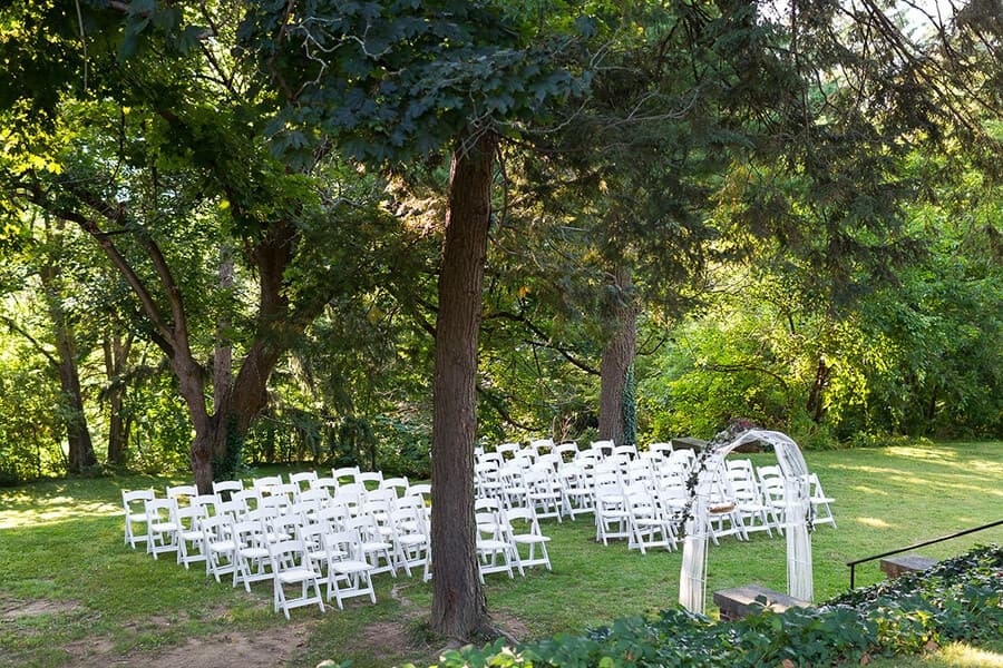 A landscaped area set up for an outdoor event, featuring rows of white chairs beneath trees.