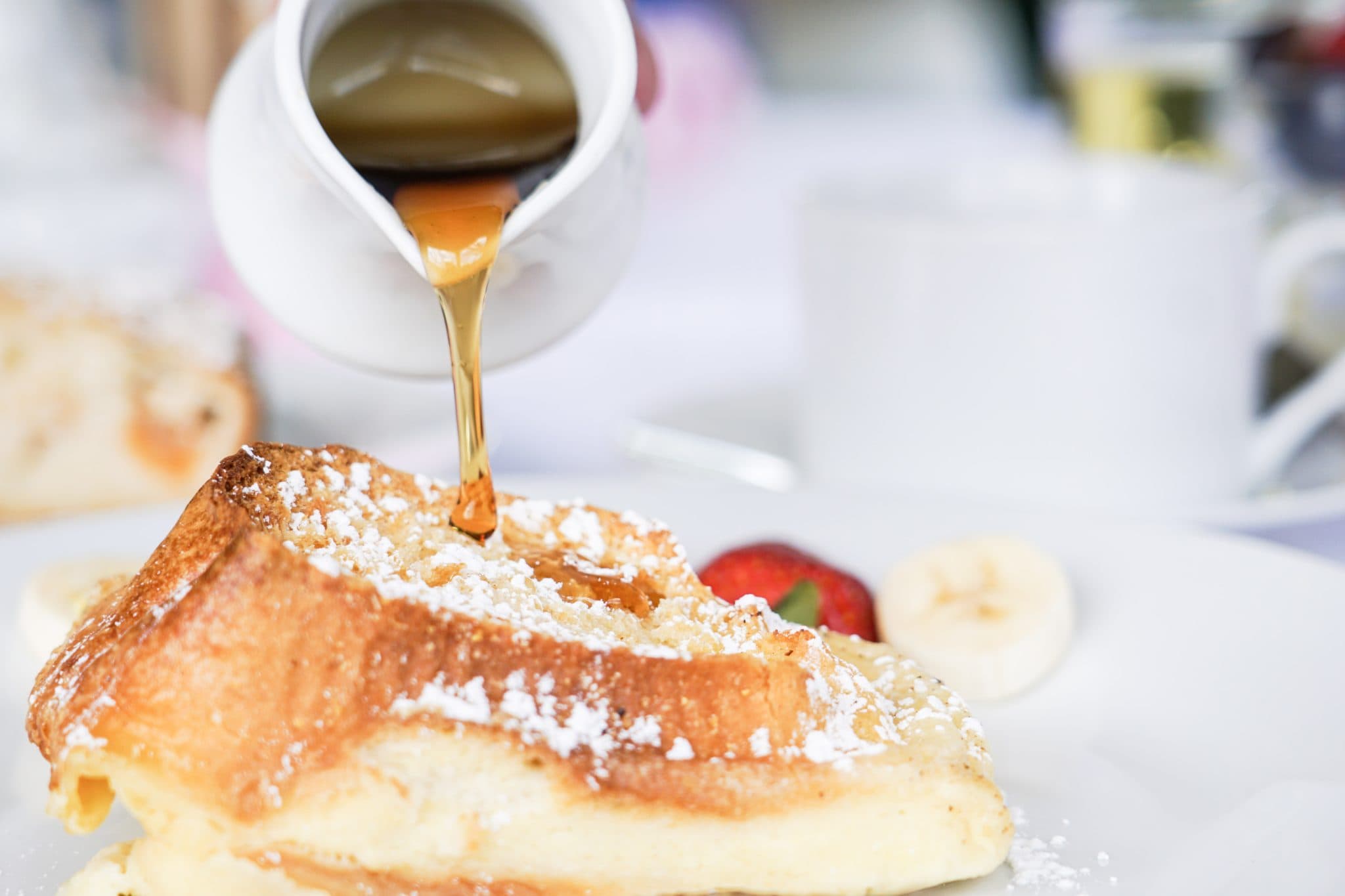 A drizzle of syrup pours over a fluffy pancake topped with powdered sugar and accompanied by fruit.