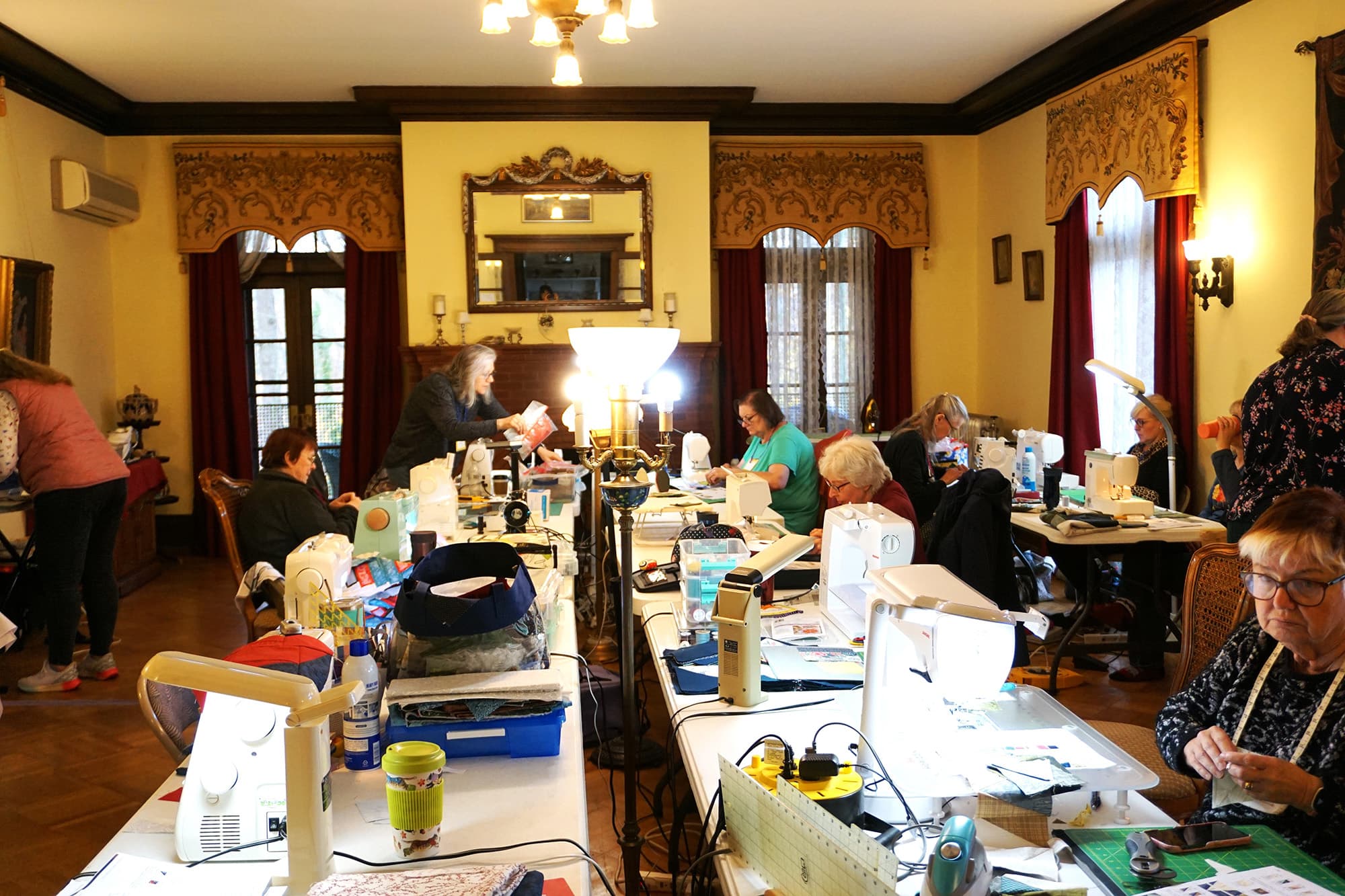 A lively sewing workshop with multiple people working at tables filled with sewing machines and materials in a well-lit room.