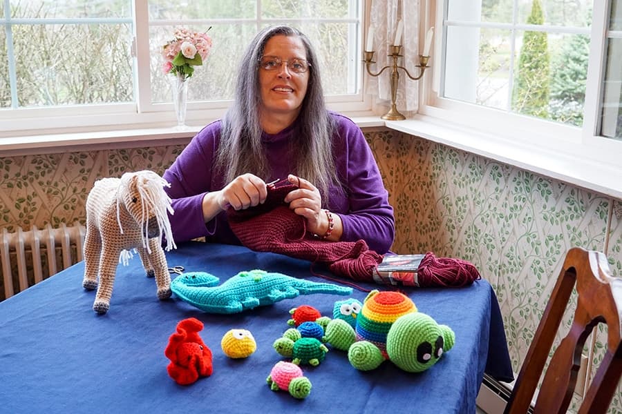 A woman with long hair sits at a table, knitting, surrounded by colorful crocheted toys.