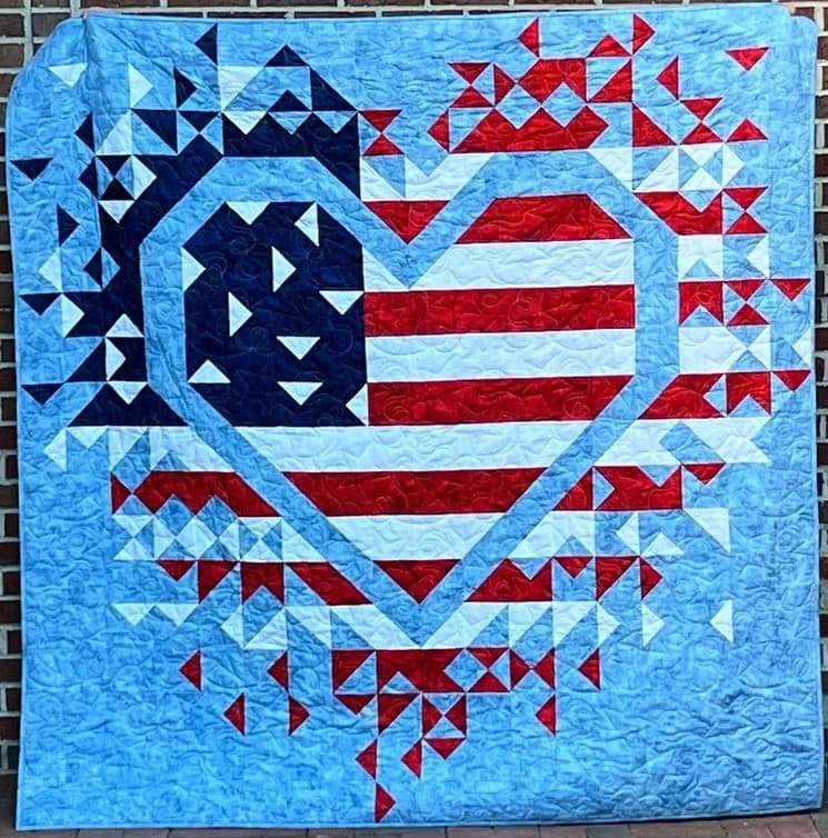 A quilt featuring a heart design with American flag colors and patterns on a blue background.