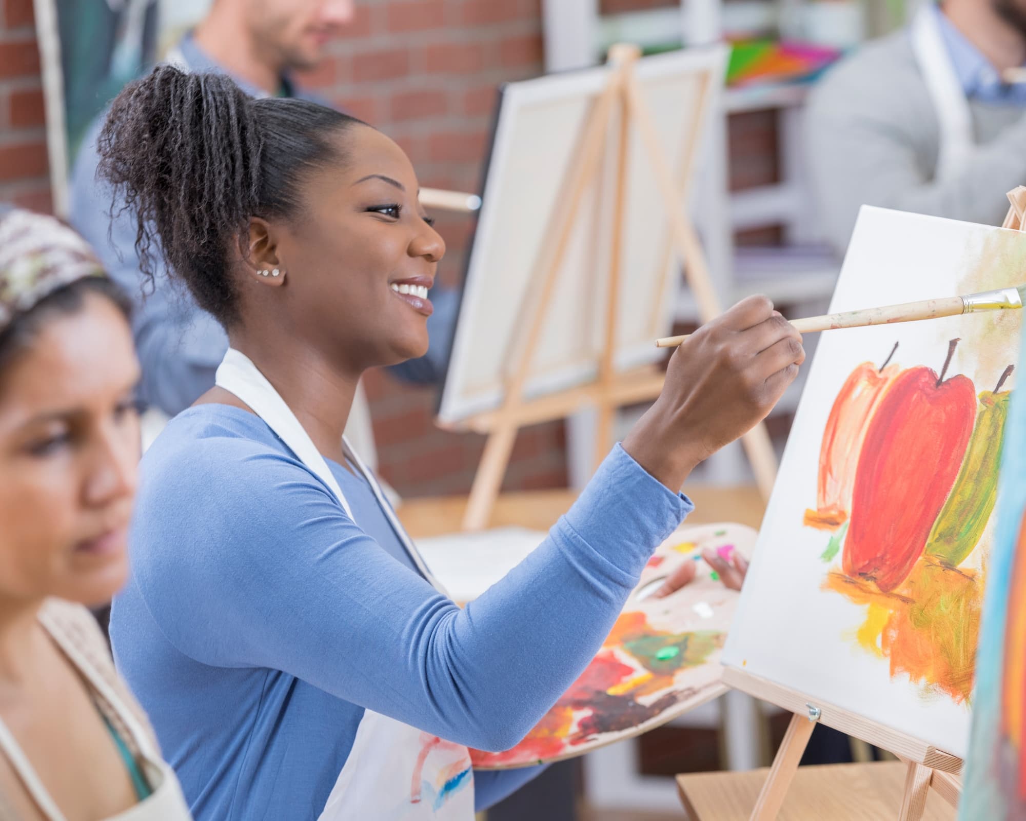 A smiling woman paints colorful peppers on an easel in an art class.