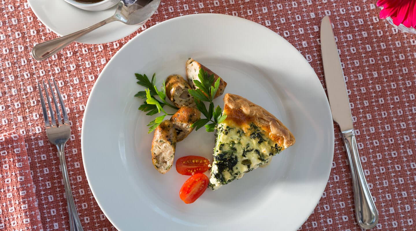 A plate featuring a slice of spinach quiche, grilled chicken, cherry tomatoes, and parsley on a patterned tablecloth.