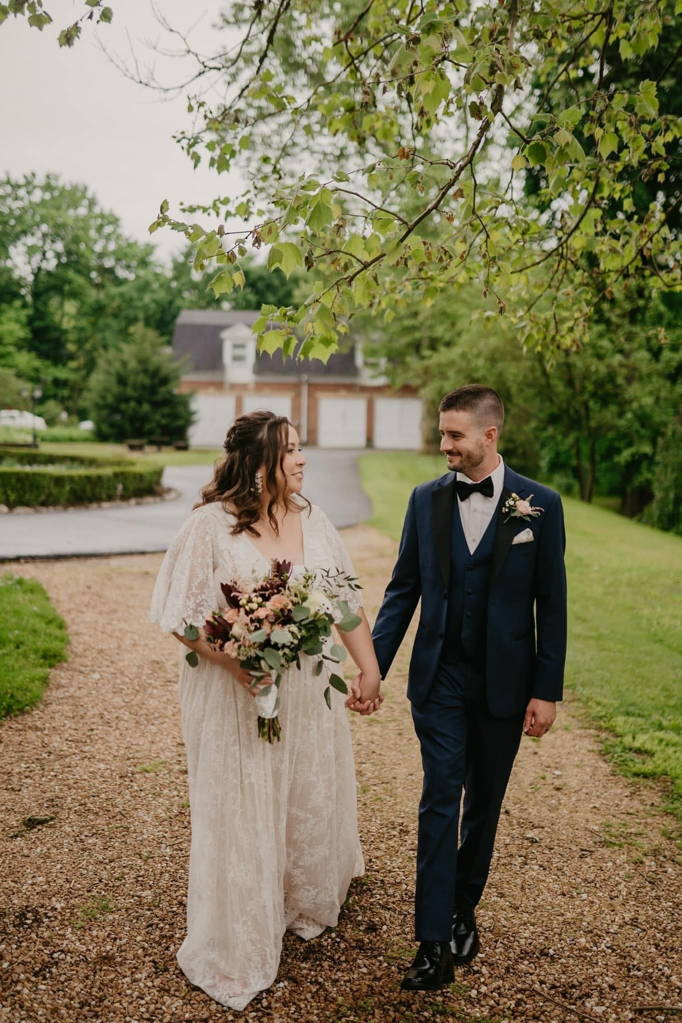 A smiling couple in wedding attire walks hand-in-hand along a gravel path surrounded by greenery.