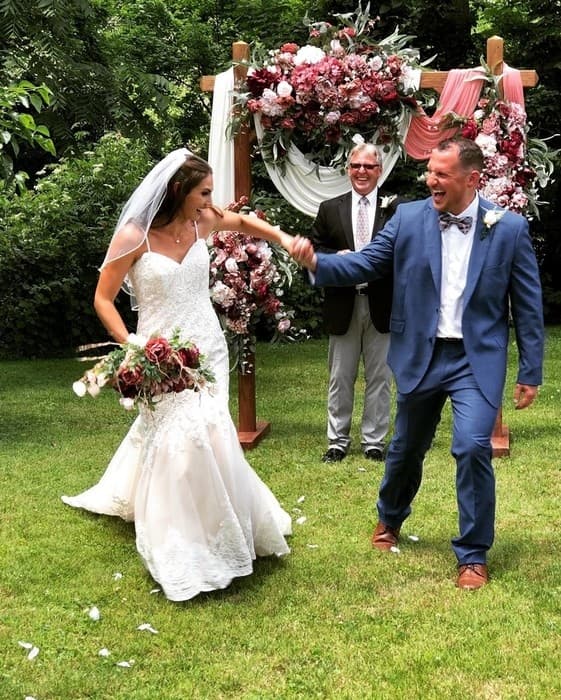 A joyful bride and groom celebrate their wedding, smiling and holding hands in front of a floral backdrop.