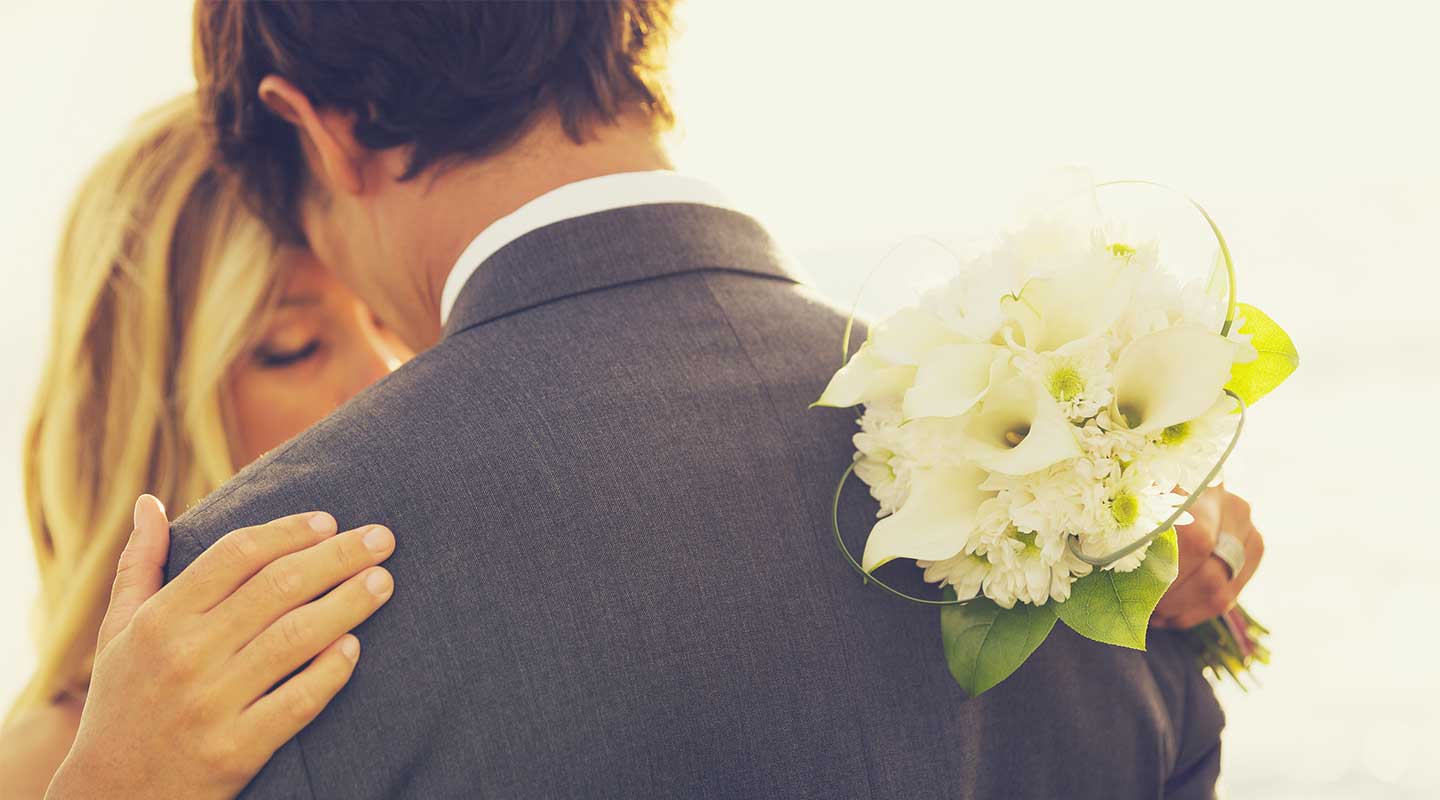 A couple embraces, with a bridal bouquet resting on the man's shoulder, against a soft, sunlit background.