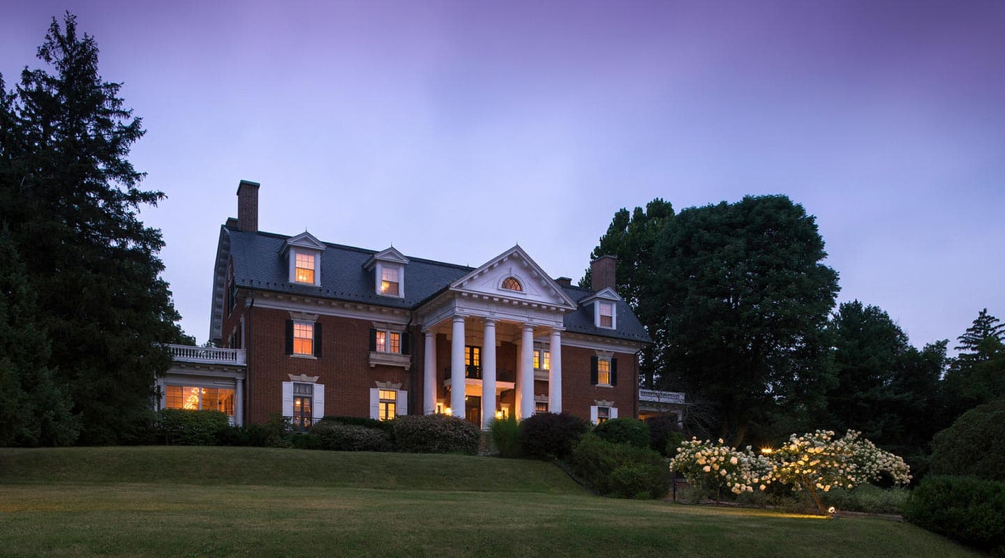 A grand brick mansion with columns, surrounded by lush greenery, illuminated at dusk.