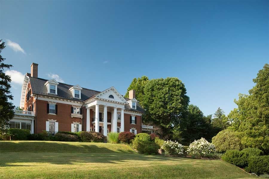 A large, historic brick house with white columns set in a lush green landscape under a clear blue sky.