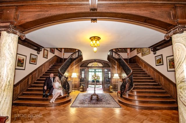 A couple sits on a staircase in an elegant foyer with wooden details and decorative artwork.