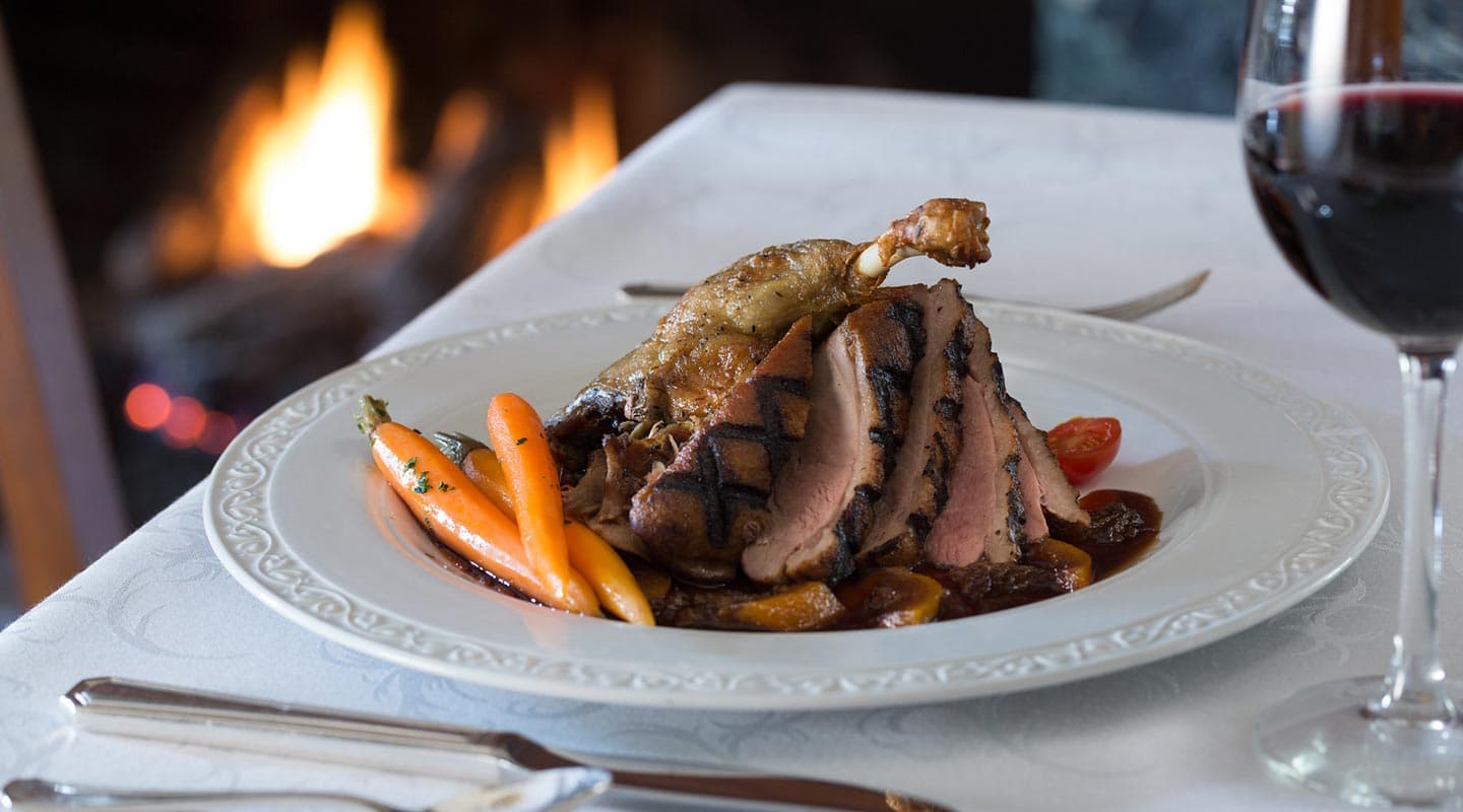 A gourmet plate of sliced grilled duck breast served with carrots and a glass of red wine by a fireplace.