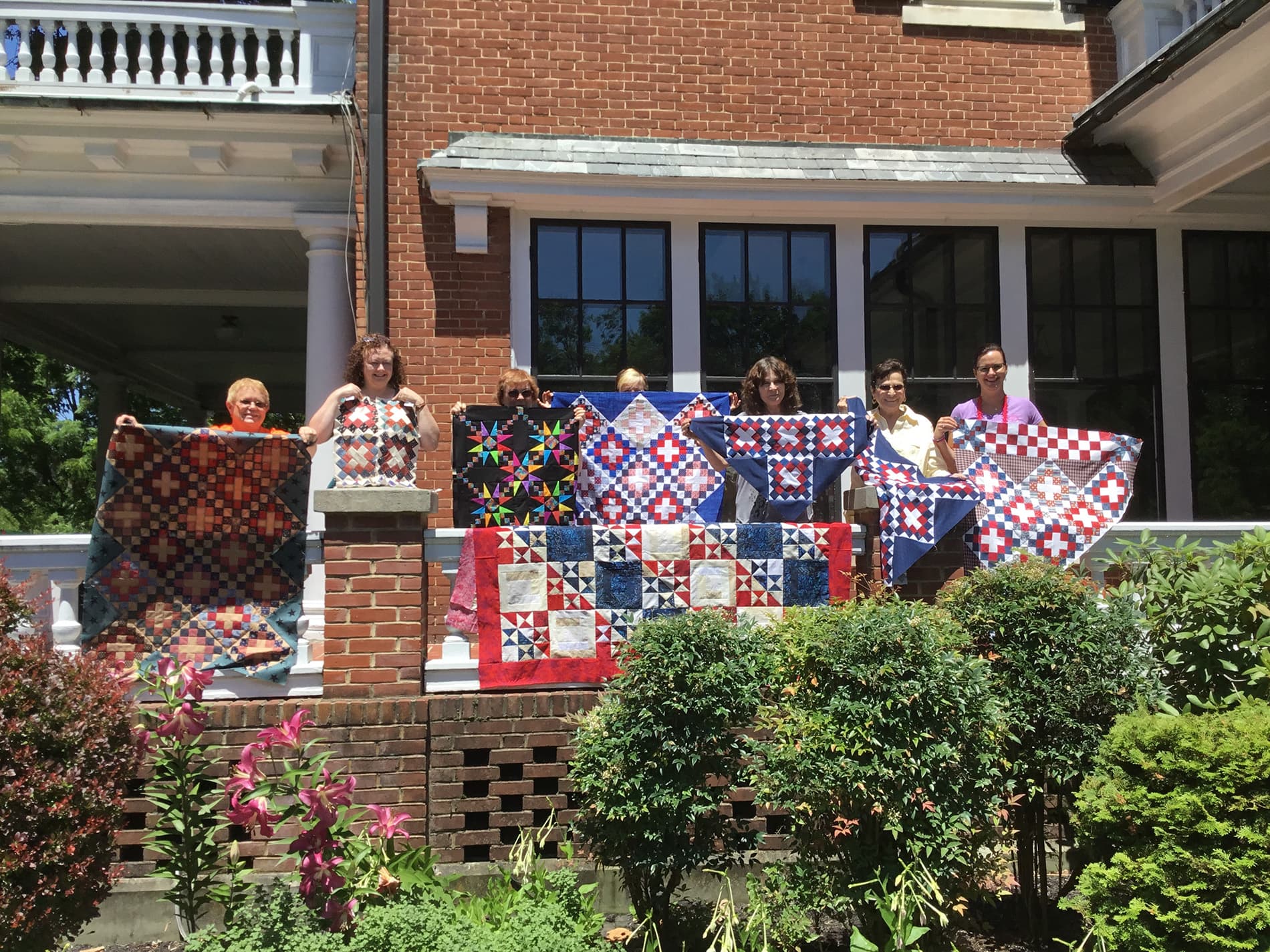 A group of women stands in front of a brick building, proudly displaying colorful quilts.