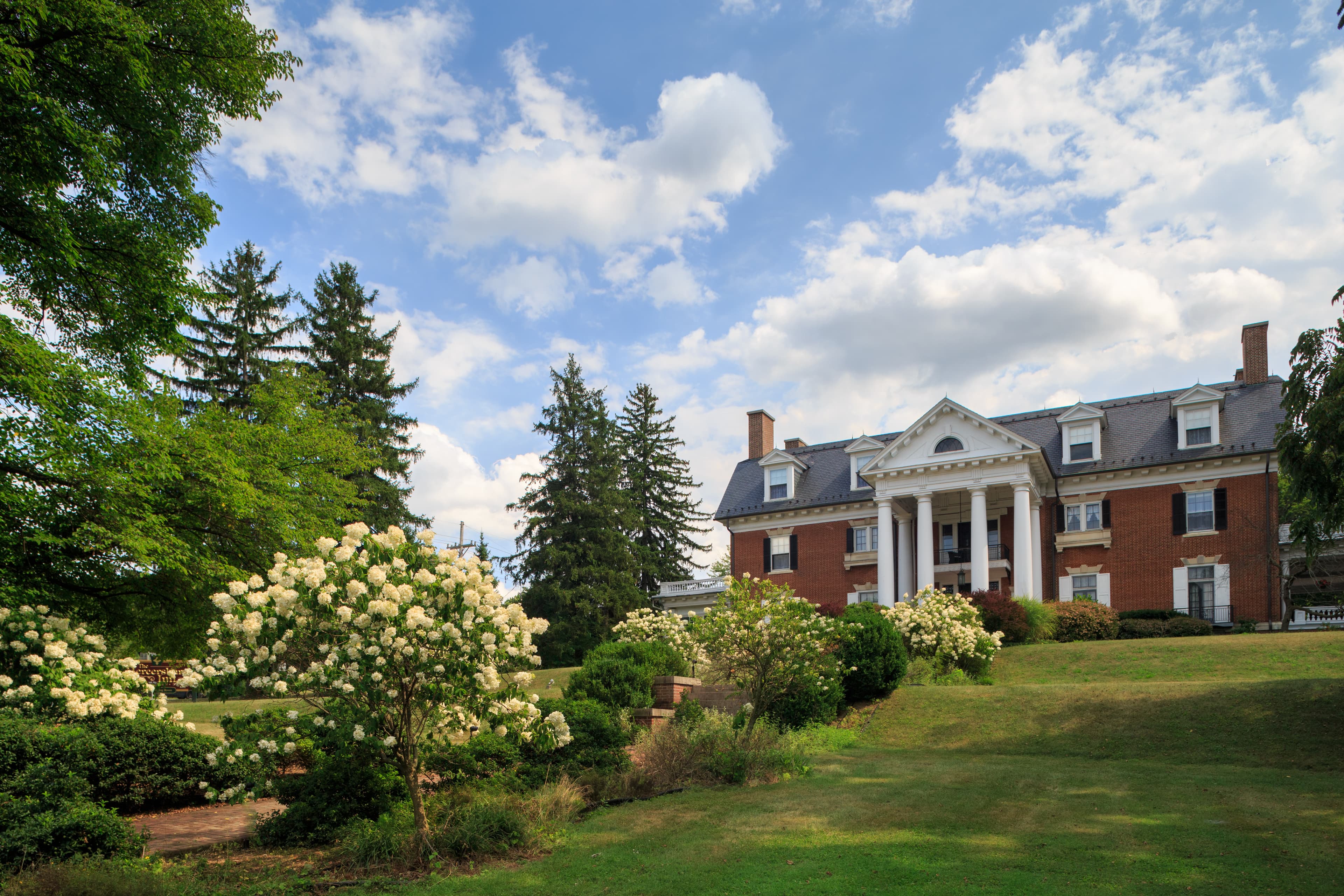 A large brick mansion with white columns is surrounded by green lawns and blooming shrubs under a partly cloudy sky.