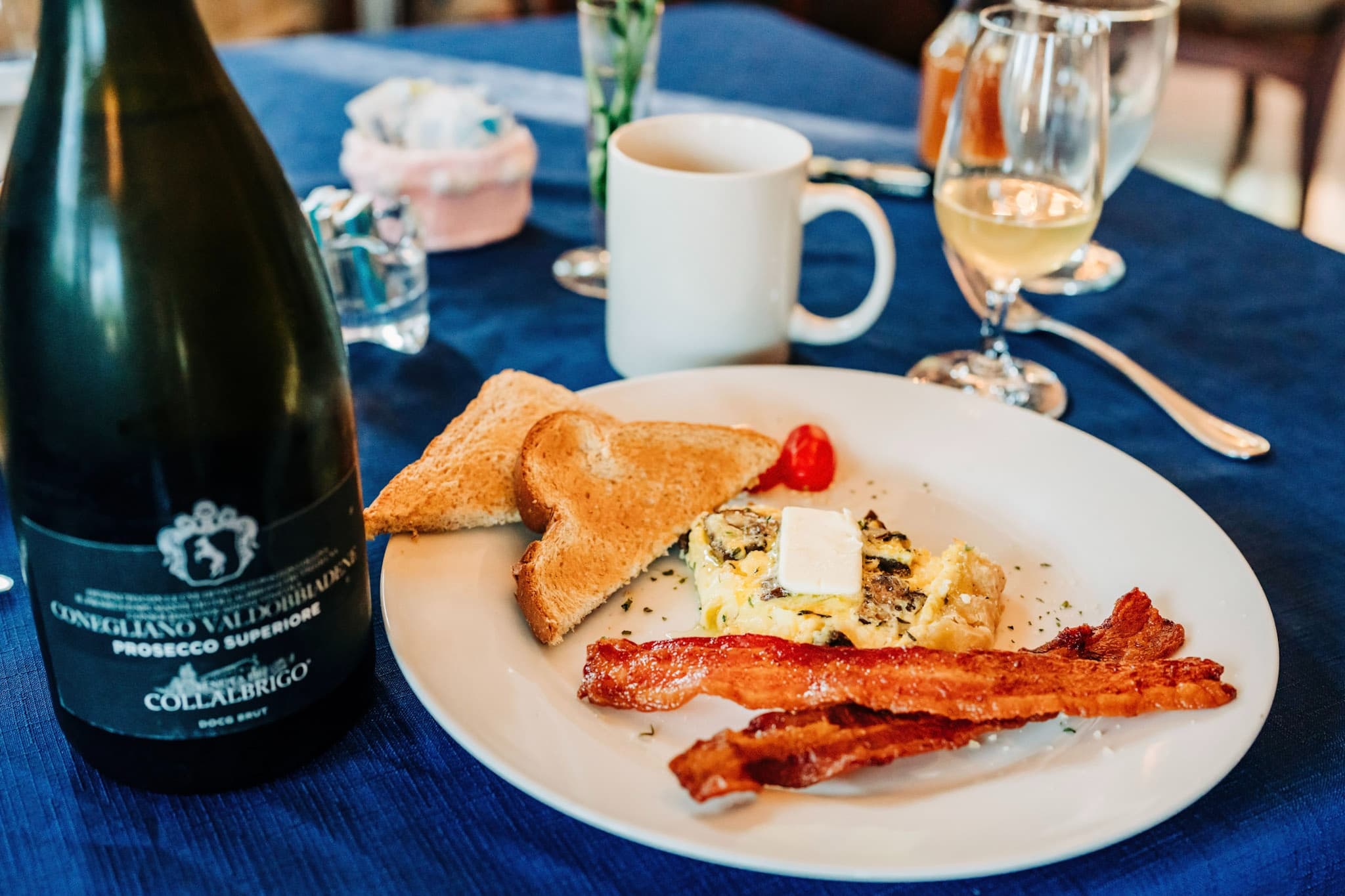 A plate with scrambled eggs, toasted bread, crispy bacon, and a cherry tomato, accompanied by a bottle of Prosecco and drinks on a blue tablecloth.