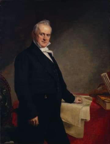 A well-dressed man stands beside a table covered with a cloth, gazing confidently at the viewer.