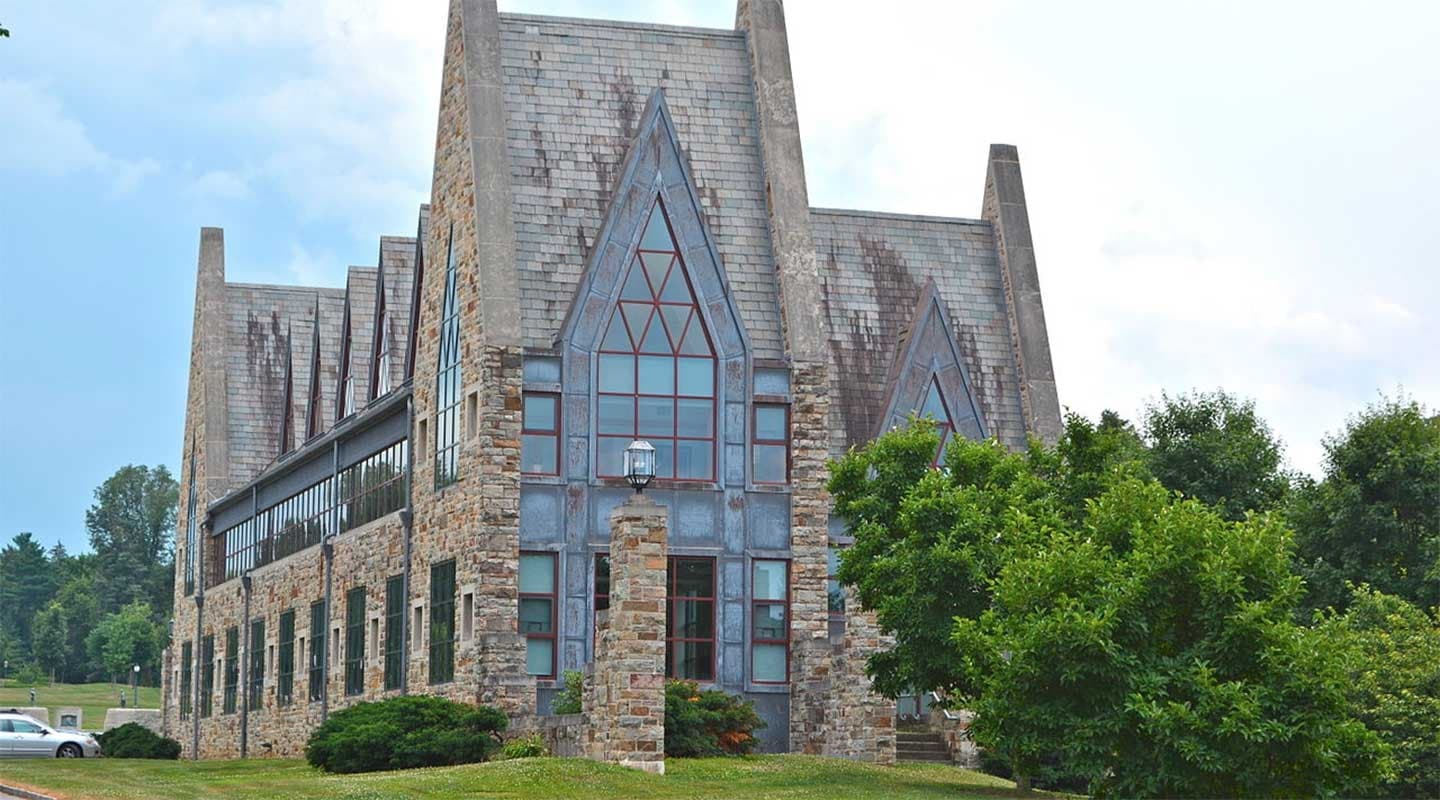 A stone building with a steep, gabled roof and large windows surrounded by greenery.