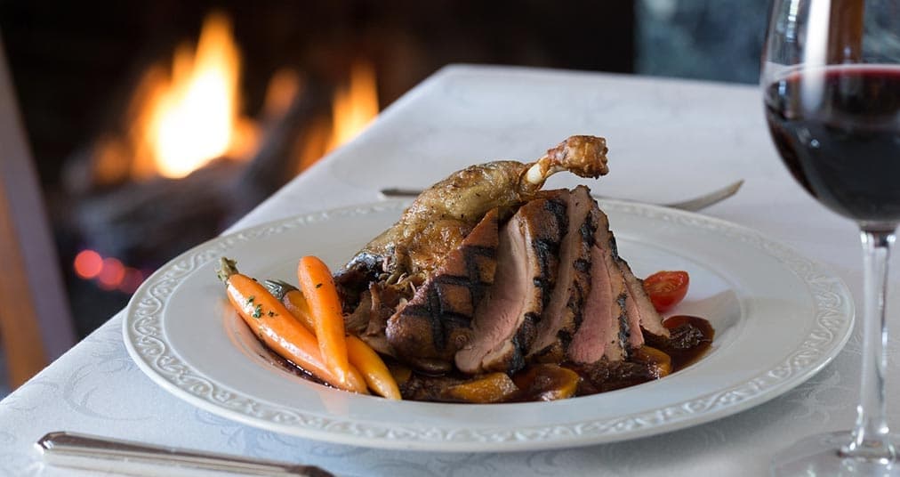 A beautifully plated portion of roasted duck with grilled vegetables, accompanied by a glass of red wine, near a glowing fireplace.