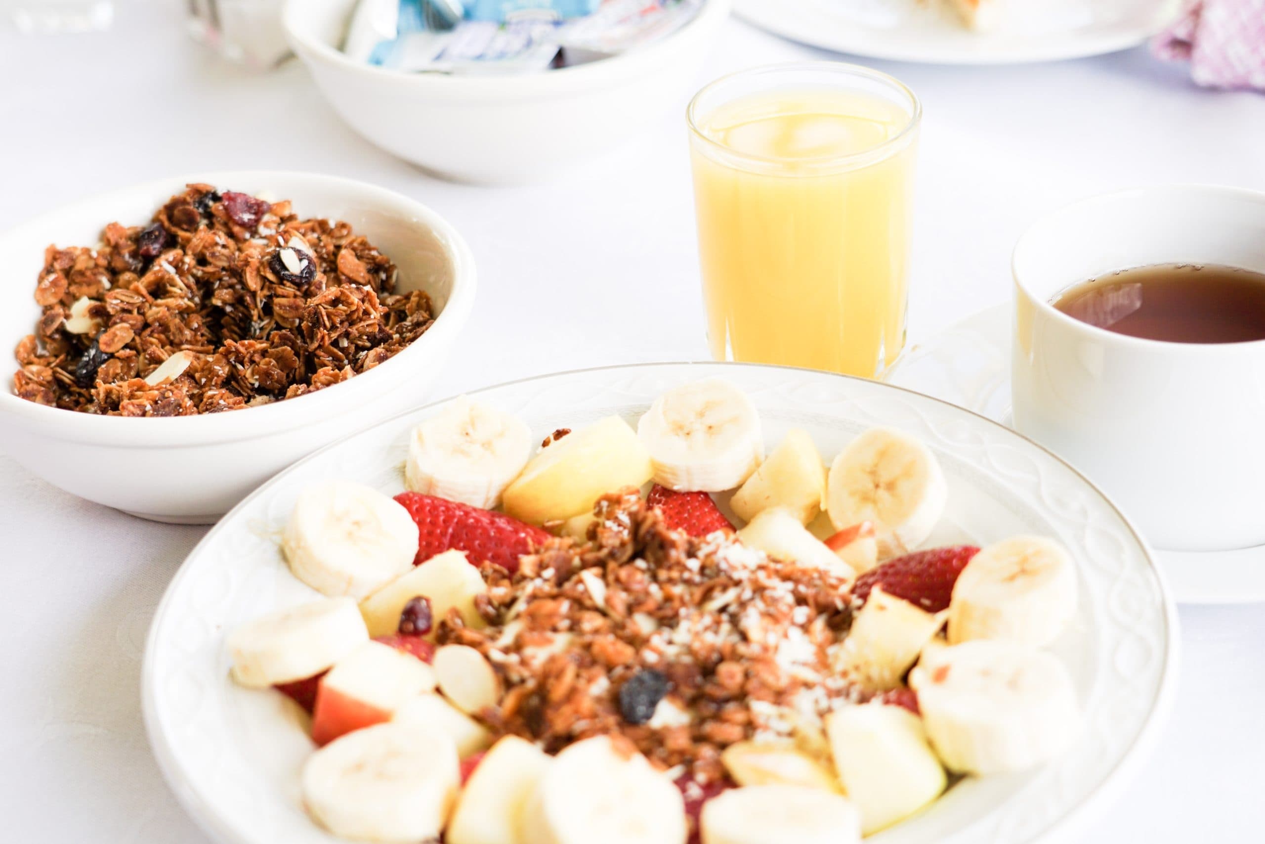 A colorful breakfast plate with sliced bananas and apples, granola in a bowl, orange juice, and a cup of tea.