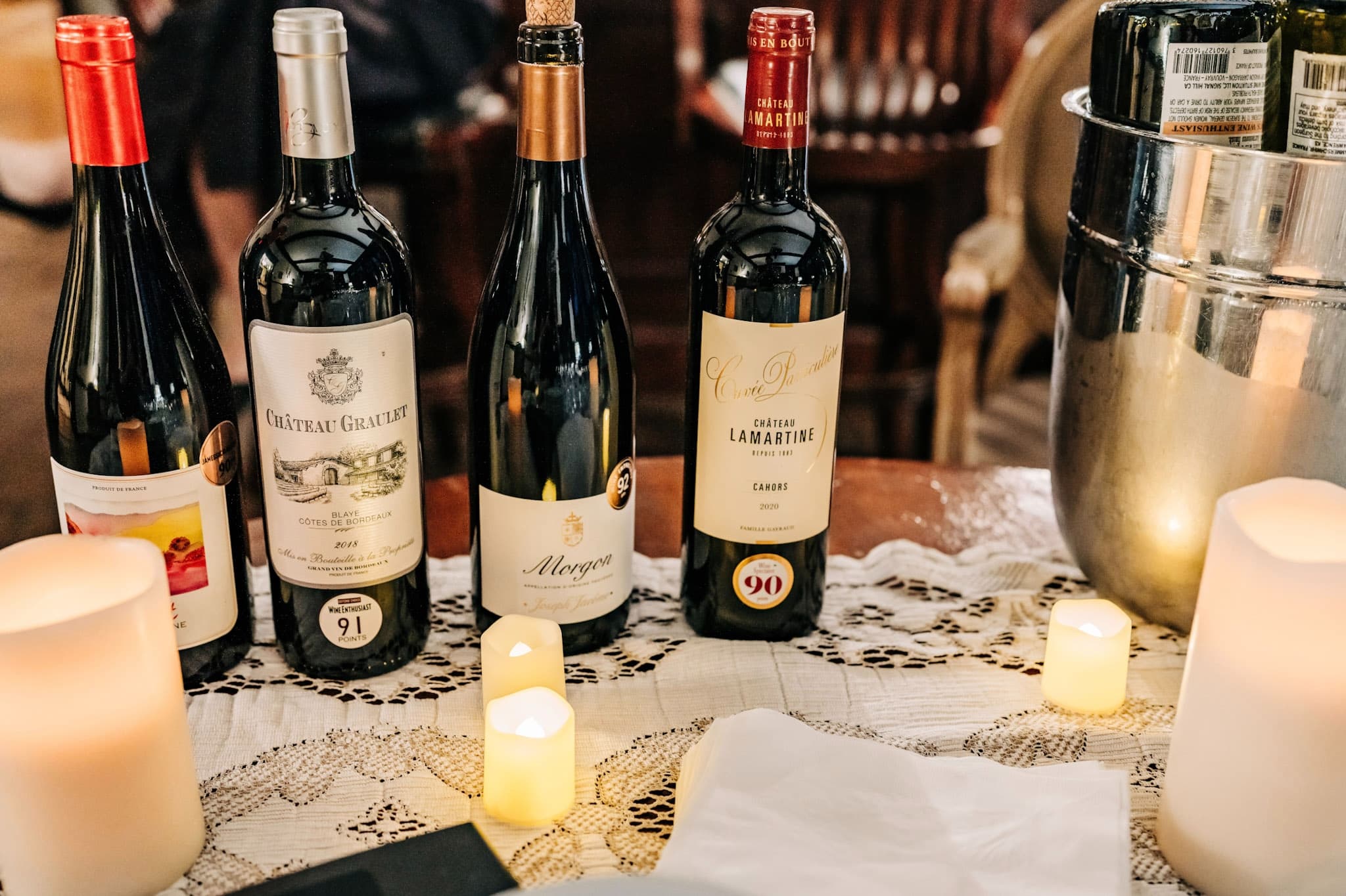A selection of red wine bottles on a lace tablecloth with flickering candles.