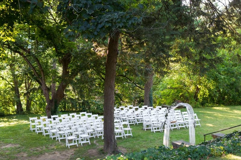 An outdoor wedding setup with white chairs arranged in rows under trees, featuring an arch at the front.